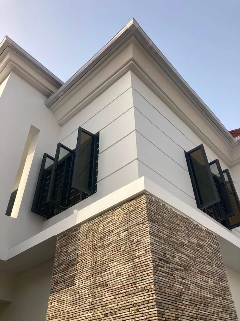 Close-up of the renovated façade showing white wall finishes, dark window screens, horizontal detailing, and textured stone cladding at the lower section.