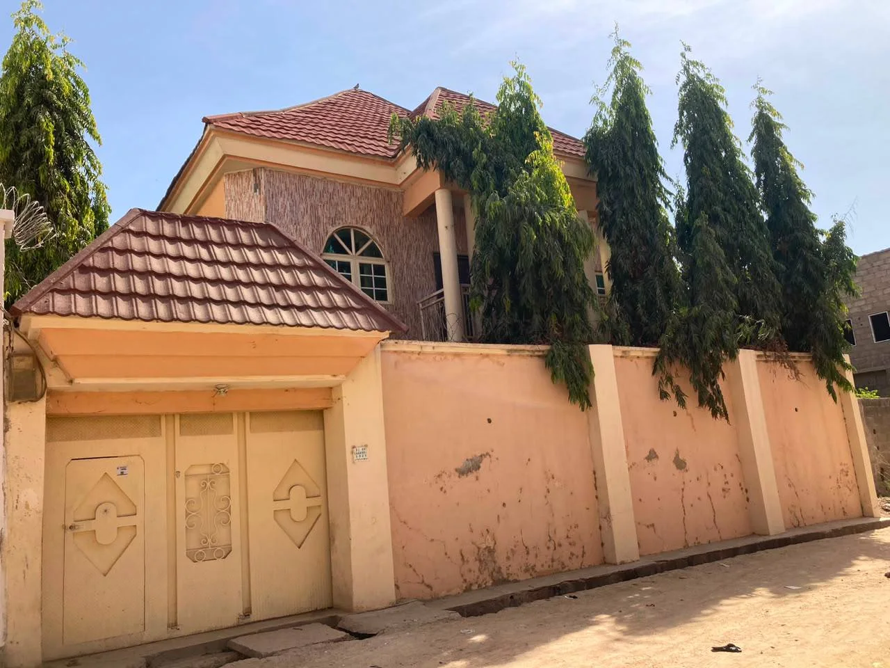 Existing residential duplex before renovation, showing the original façade, tiled roof, boundary wall, entrance gate, and mature trees screening the property.