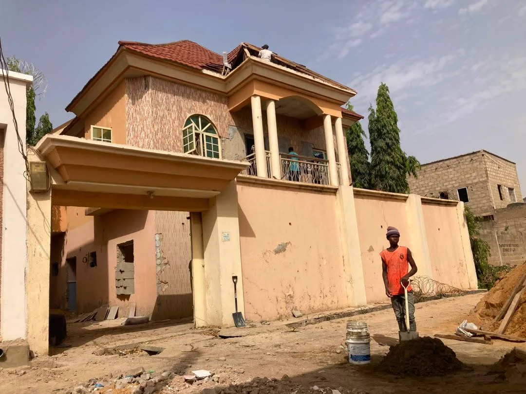 Existing residential duplex before renovation, showing the original façade, boundary wall, entrance canopy, tiled roof, and site preparation works.